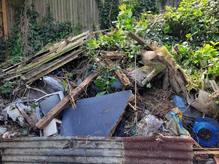 Overgrown corner of a garden piled with old timber, carpet and junk before clearance